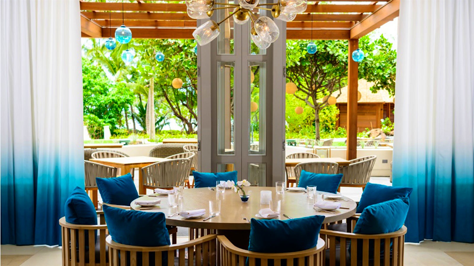 A private dining room at Noe, outside under a wooden pergola, with light-brown wooden chairs and blue cushions, and glass lighting fixtures hanging overhead